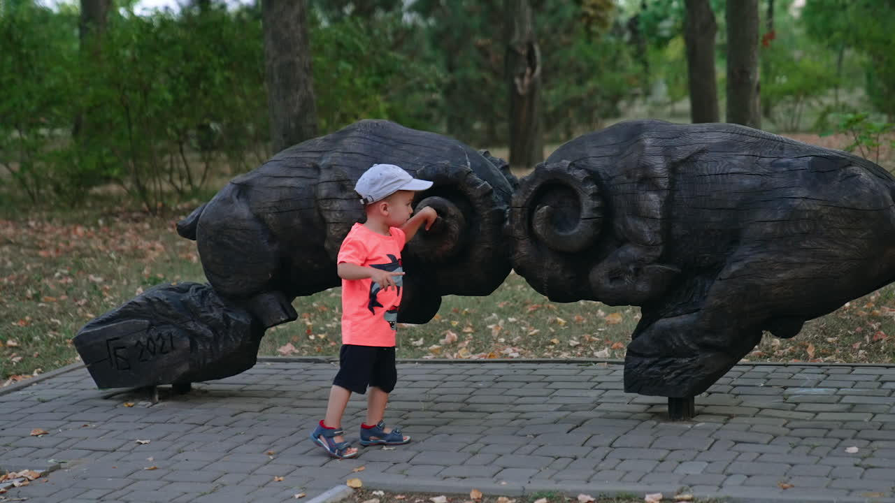Thoughtful Caucasian baby in cap stands in the park near the wooden sculptures. Cute kid touching the figures gently.