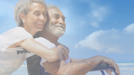 animation of glowing light over smiling senior man in wheelchair with his wife by seaside