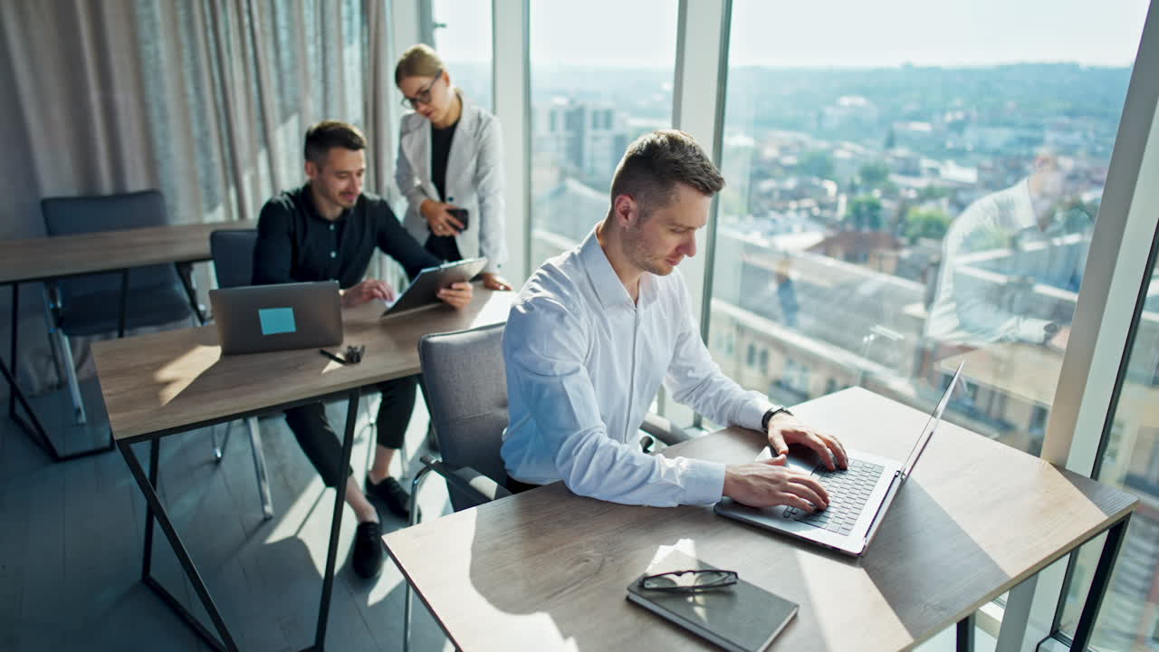 Young office team working in the office. Man in white shirt sitting at front desk working at laptop. Male in black shirt behind talks to female colleague standing next to him discussing documents.