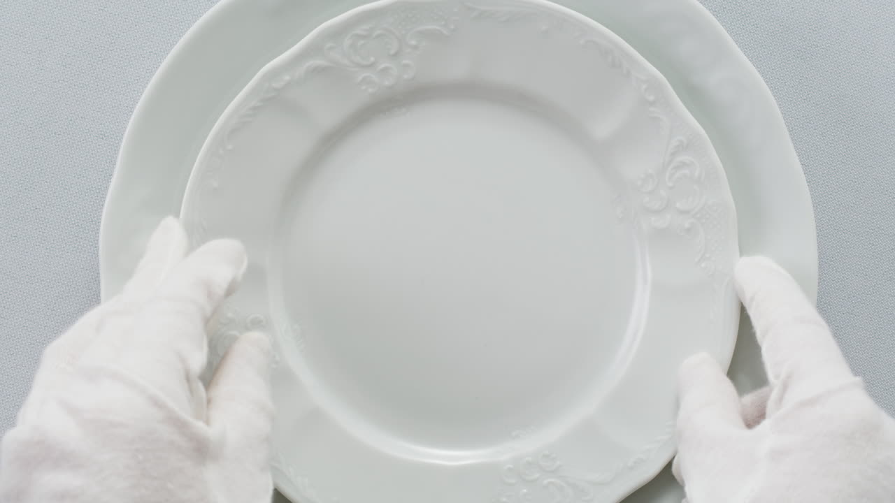 Professional table setting with white gloves. Plates, fork, knife, and glass arranged on a white table.