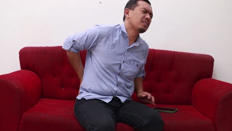 unhappy middle-age asian man suffering from backache while sitting on the sofa at home