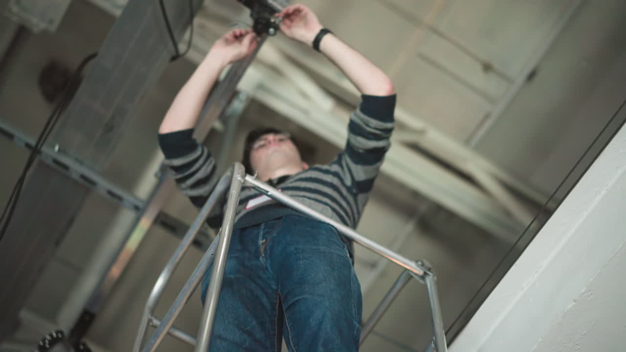 Worker using tools to install equipment on elevated platform in broadcast studio, high-angle shot showing ceiling infrastructure and cables, focused on installation process