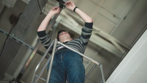 worker using tools to install equipment on elevated platform in broadcast studio, high-angle shot showing ceiling infrastructure and cables, focused on installation process