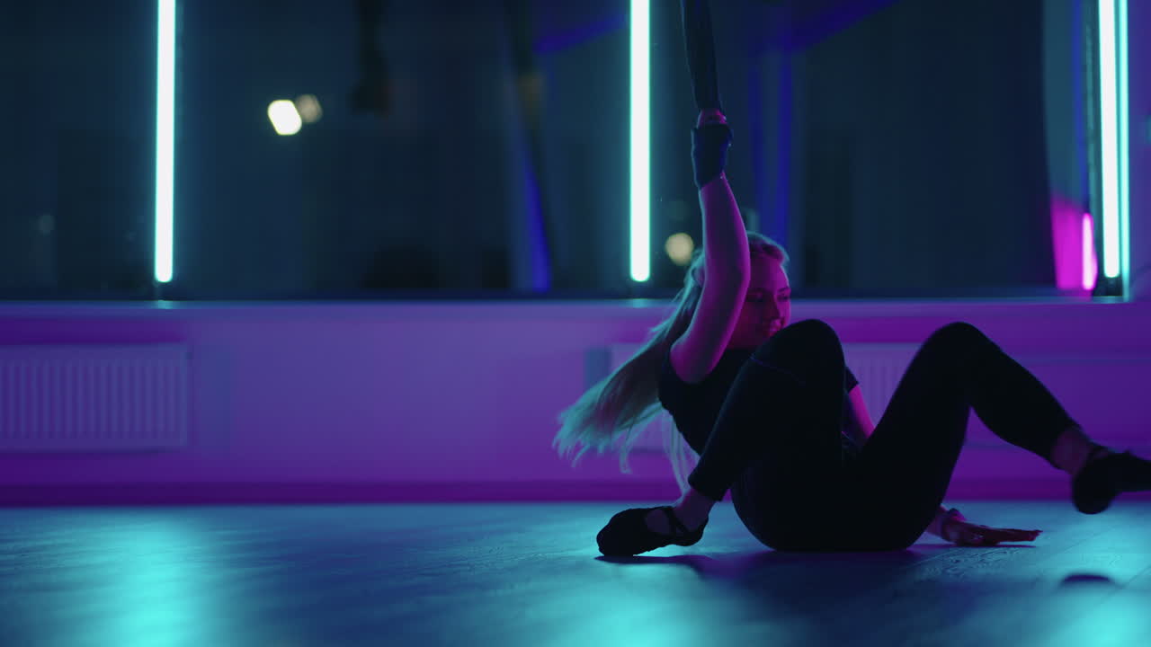 A woman does yoga and stretching on a hanging hammock in neon light. A woman is flying in a hammock in the studio doing stretching and body exercises. Exercises in the air