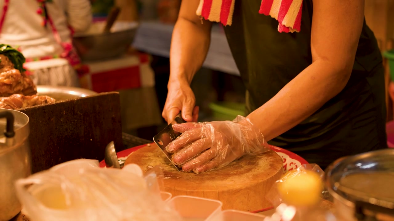 Primo piano delle mani di uno chef che affettano carne su un tagliere di legno sotto la calda illuminazione di un mercato notturno