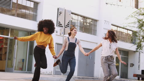 tres amigas milenarias tomadas de la mano y riendo mientras corren a través de un cruce de peatones