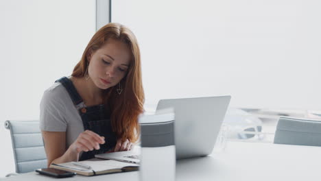 Joven-Empresaria-Vestida-Informalmente-Trabajando-En-Una-Computadora-Portátil-En-Una-Oficina-Moderna-Atendiendo-Una-Llamada-Por-Teléfono-Móvil