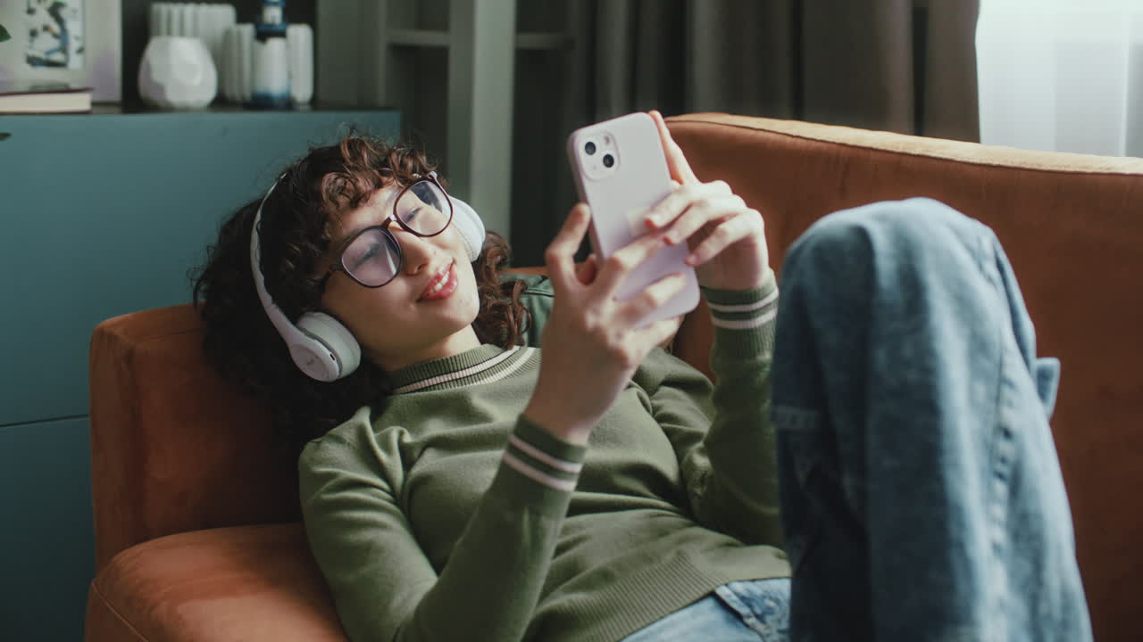 Young Smiling Woman in Headphones Lying on Sofa, Enjoying Music and Using Phone