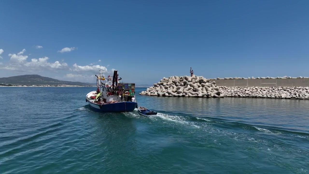 Veduta aerea di un peschereccio che naviga accanto a un lungo frangiflutti mediterraneo nel Marocco settentrionale, lasciando una scia visibile attraverso le calme acque blu del porto.