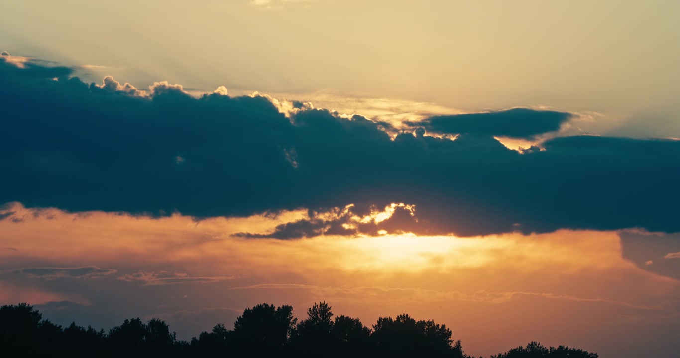 Golden sunset shining through dramatic storm clouds above a silhouetted tree line. Warm evening light and layered clouds create an atmospheric weather landscape.