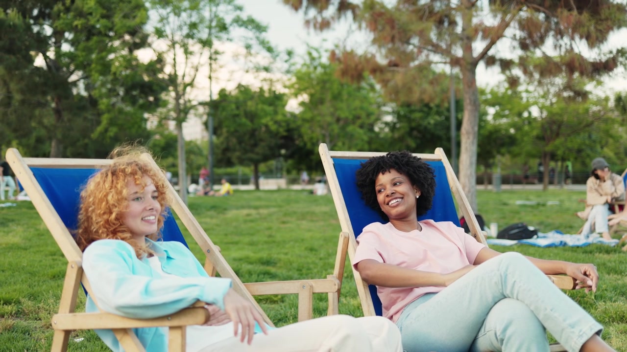 Two Diverse Young Women Relaxing and Talking in Deck Chairs in a Park