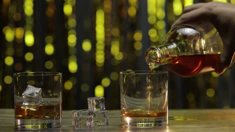 Pouring-of-golden-whiskey,-cognac-or-brandy-from-bottle-into-glass-with-ice-cubes.-Shiny-background