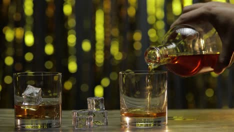 pouring of golden whiskey, cognac or brandy from bottle into glass with ice cubes. shiny background