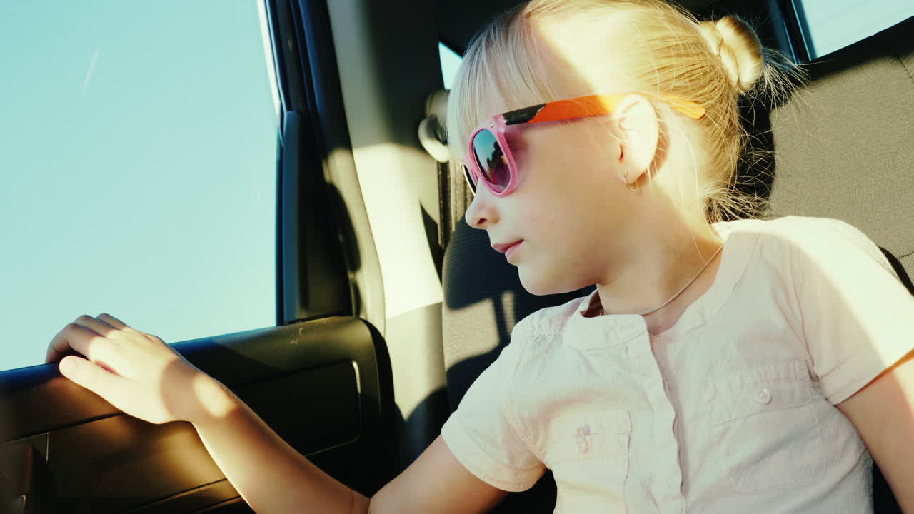 una niña de 6 años con gafas rosas protectoras contra el sol viaja en el asiento trasero del automóvil que está sujeto