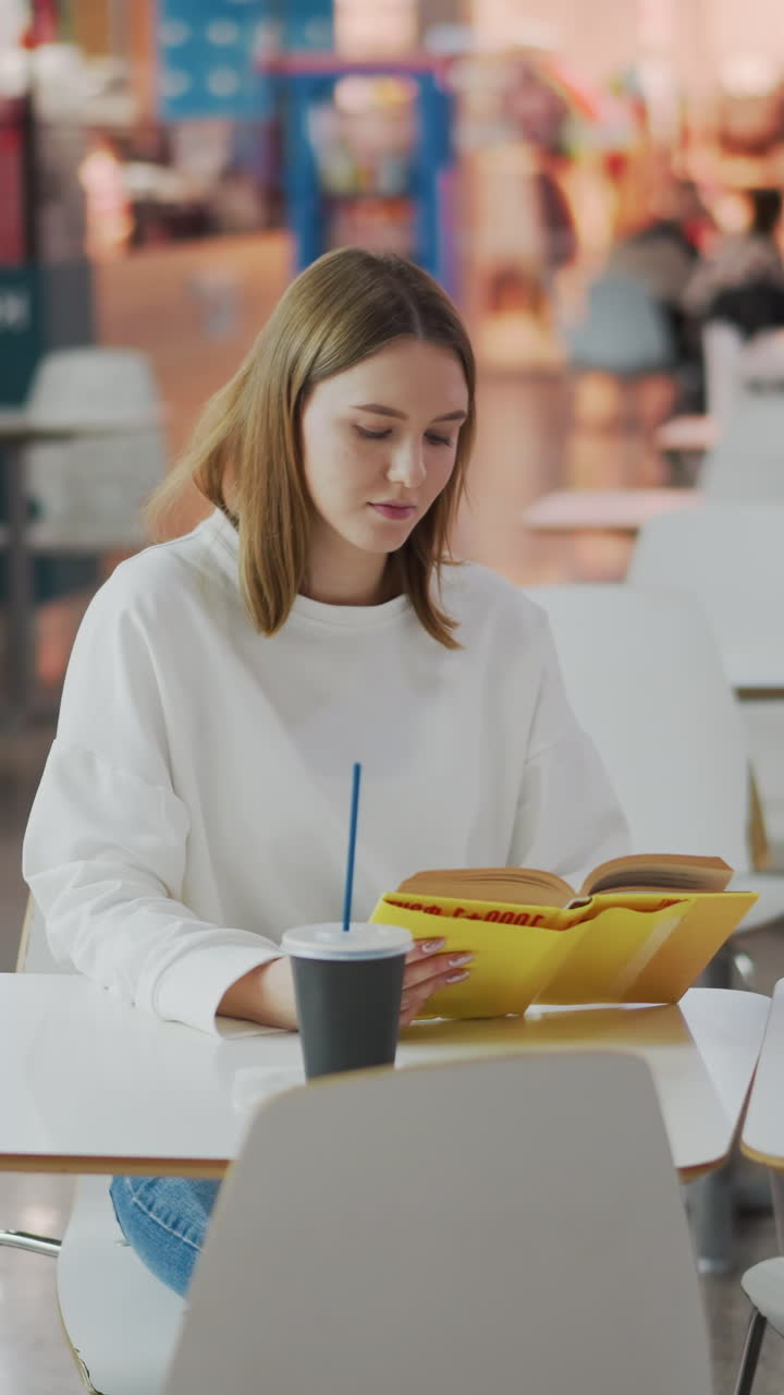 Young woman seated at table reading book in vibrant mall surrounded by shopping bags, soft lighting, and colorful background with other shoppers visible in distant seating