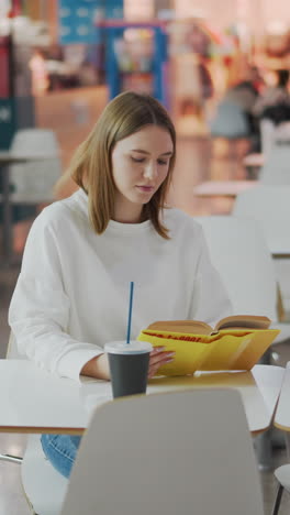 mujer joven sentada en una mesa leyendo un libro en un vibrante centro comercial rodeada de bolsas de compras, iluminación suave y fondo colorido con otros compradores visibles en asientos distantes