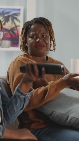 indian woman looks for interesting tv channel while african american girl friend drinks tea. best female friends enjoy watching program together at home