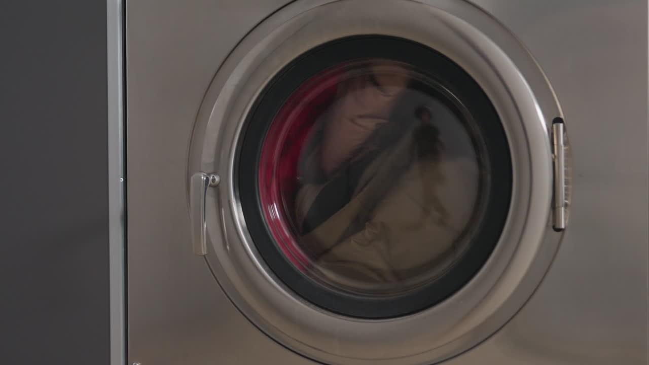 Front load industrial washer in operation with mixed clothes swirling inside drum, stainless door marked 2, laundromat scene, spinning cycle, clear view of red garment motion and wash