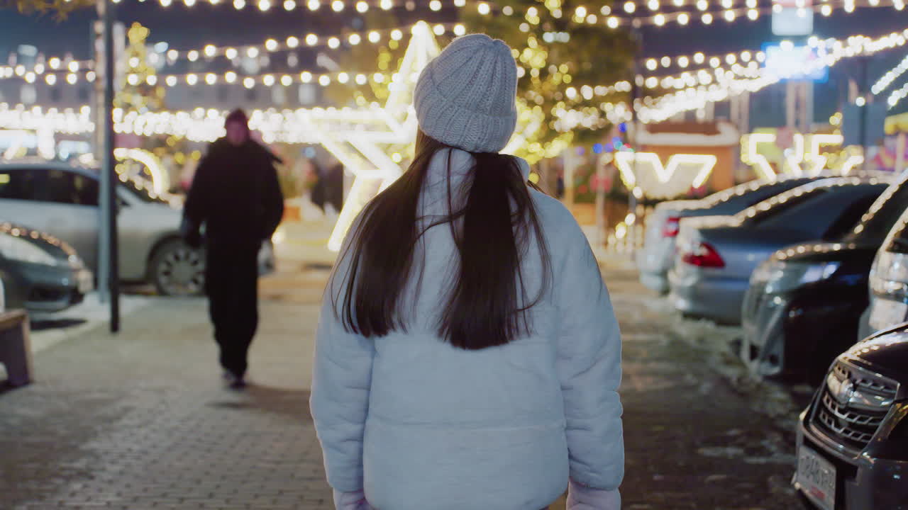 Rear view of woman in cozy winter outfit walking through a beautifully decorated city at night, festive lights create a warm bokeh background, with parked cars and people