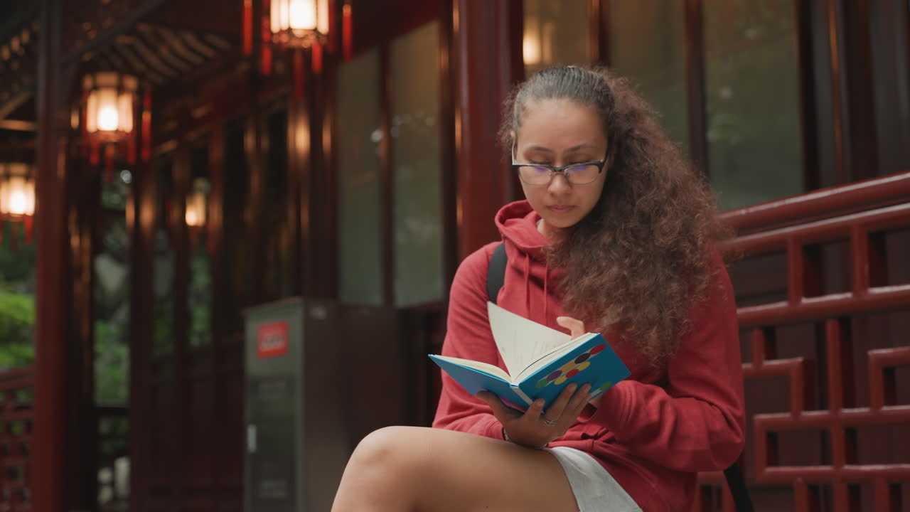Jeune femme lisant un carnet coloré en extérieur, cheveux bouclés et lunettes, portant un sweat à capuche rouge, assise sur un banc en bois près d'un pavillon éclairé par des lanternes, alternant appel téléphonique et prise de notes, bokeh de soirée.