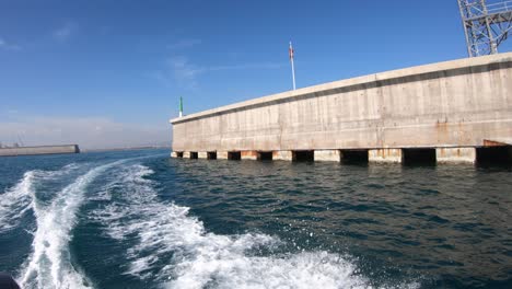 Lancha-Saliendo-Del-Puerto-De-Castellon,-El-Faro-Y-La-Muralla-Con-La-Estela-Del-Motor-En-El-Agua-De-Mar