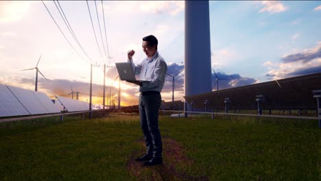 full body side view of an asian male professional worker standing with his laptop with solar panel and wind turbines, he raises his fist up with screaming goal after typed on the laptop