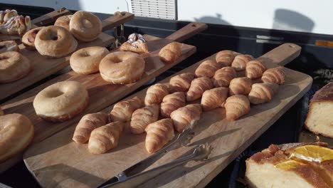 wooden serving board gradually filling with gourmet pastries, donuts, and croissants, displaying increasing variety of delectable bakery treats under soft sunlight
