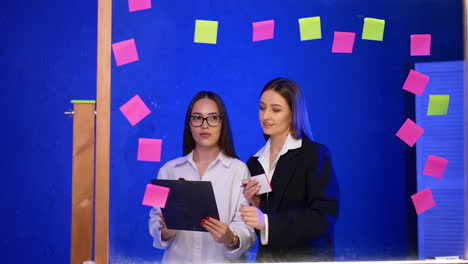 female colleagues work out ideas near the glass wall. women look at the clipboard and on the stickers put on the board.