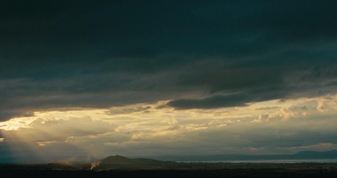 Dark storm clouds with dramatic sunbeams breaking through over a distant landscape. Moody weather, layered clouds, and atmospheric light create a powerful natural scene.