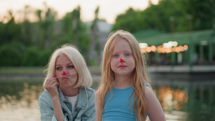 retrato de mãe e filha sorrindo à beira do lago ao pôr do sol pintando os rostos com tinta colorida usando pincéis, capturando um momento de arte divertido, respingos coloridos na pele, conexão alegre