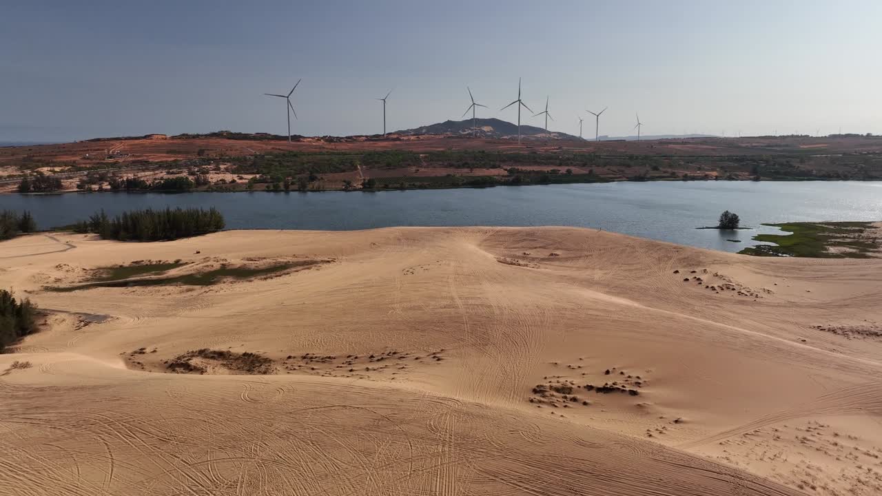 Aerial view of vast sand dunes, tranquil lake, and wind turbines on the horizon in Vietnam, blending natural desert-like landscapes with renewable energy and serene rural scenery