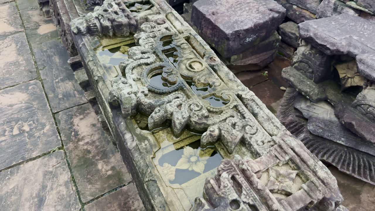 A slow push-in reveals a weathered carved stone fragment, its surface etched with ornate floral and scroll motifs, rainwater resting in its grooves, echoing ancient artistry and timeless devotion