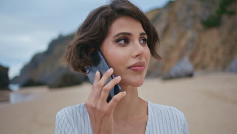 relajada morena hablando por teléfono móvil en la playa de cerca. mujer tranquila caminando por la orilla