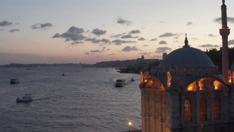 Mezquita-Ortakoy-Bellamente-Iluminada-Con-Luz-Amarilla-En-La-Orilla-Del-Río-Bósforo-Que-Revela-Una-Increíble-Puesta-De-Sol-Sobre-Estambul,-Toma-Aérea-De-Establecimiento