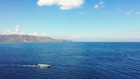 Touring-boat-with-tourists-sailing-across-deep-blue-sea,-approaching-to-tropical-island-bay-on-a-bright-sky-background-with-white-clouds-in-Bali