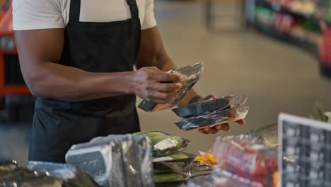 fotografía en primer plano de un hombre negro con un delantal negro y una camiseta blanca colocando mercancías en un estante en una tienda de comestibles moderna. clasificación y preparación de mercancías expuestas para los clientes en grandes tiendas