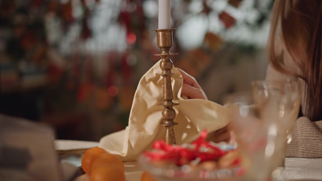 Hand polishing brass candlestick on table, bowl of red berries and citrus at side, vintage ornament backdrop, domestic ritual of care, gentle motion and mindful restoration, warm holiday ambience