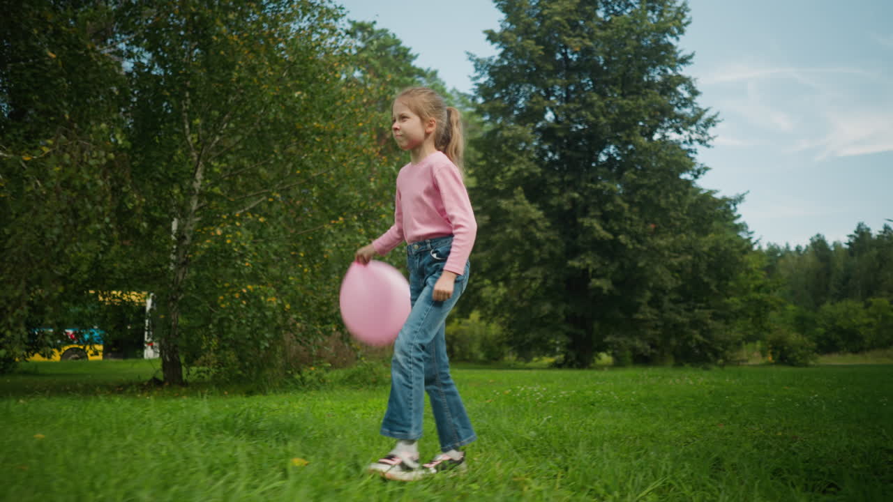 Cute girl holding pink balloon walking joyfully on grassy field surrounded by lush green trees that partially cover parked cars in background under blue sky with scattered clouds