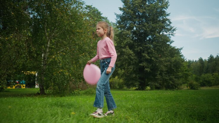 Cute girl holding pink balloon walking joyfully on grassy field surrounded by lush green trees that partially cover parked cars in background under blue sky with scattered clouds