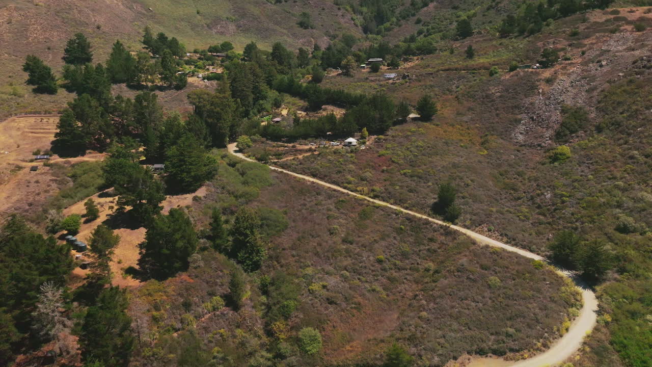 Wavy road through the rocky landscape leading to the houses among the green fir-trees. Mountains at backdrop. Top view.