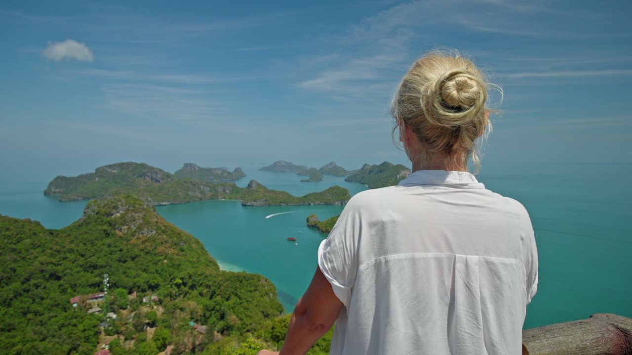 Mulher de meia-idade em pé em um mirante de madeira, segurando um chapéu de palha e contemplando o mar azul-turquesa e as ilhas tropicais do Parque Marinho de Ang Thong, um momento de viagem tranquilo na Tailândia. Filmagem em 4K de alta qualidade.