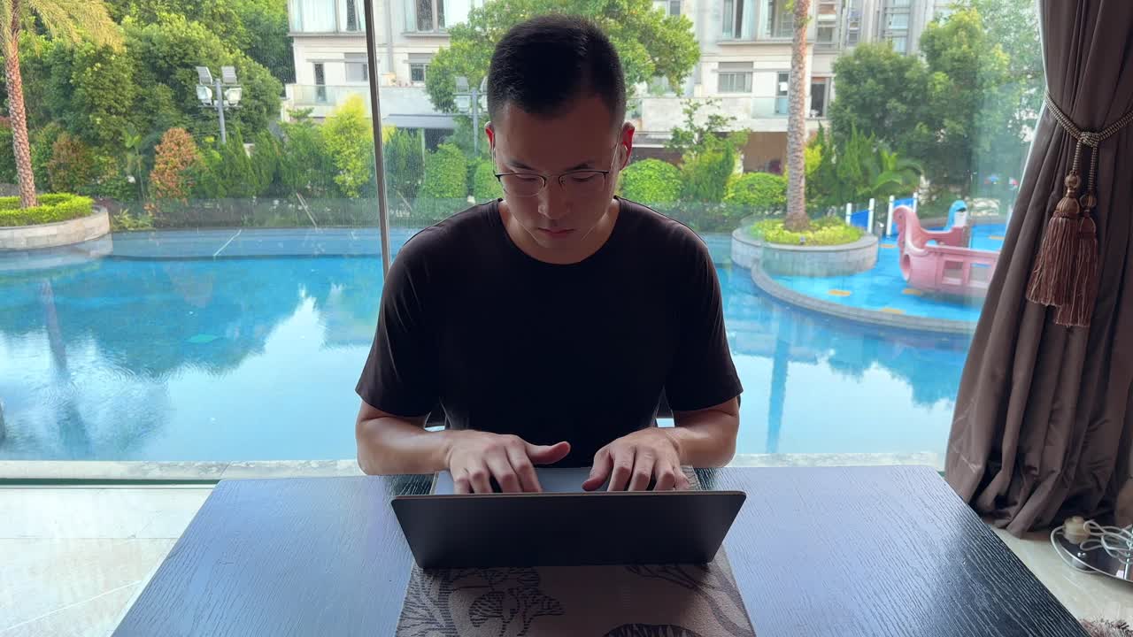 Asian Millennial Man Working Remotely at Home on Laptop in his Luxurious Home Overlooking Pool