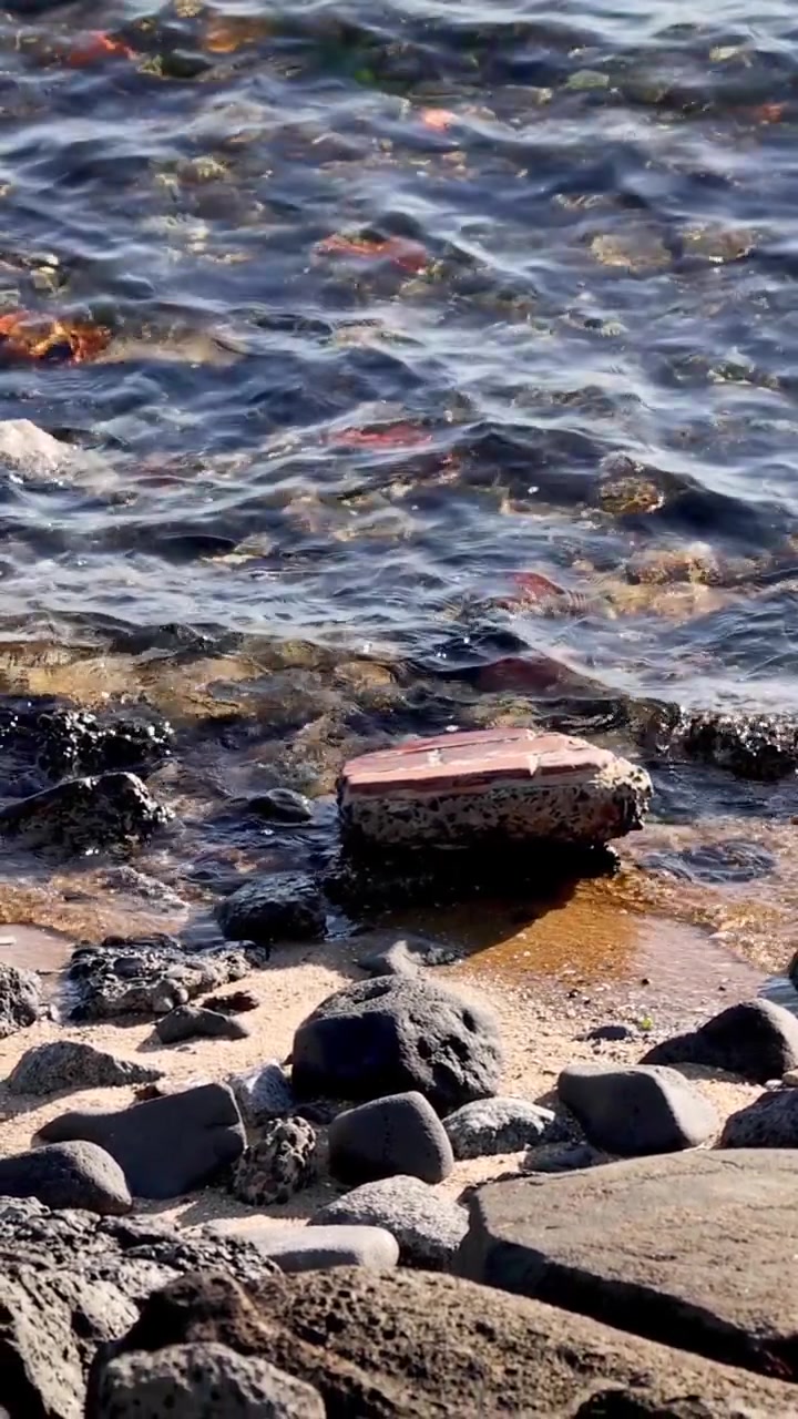 Primo piano di acqua oceanica limpida che si infrange su rocce scure e ciottoli su una spiaggia luminosa.