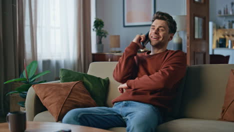 Positive hipster speaking mobile phone at home weekend. Happy man laughing couch