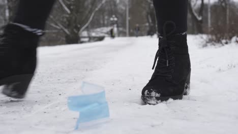 woman's feet in black boots, throws used mask on snowy ground in winter, close up