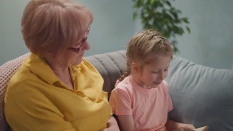 Senior-blonde-lady-sits-by-little-girl-reading-book-at-home