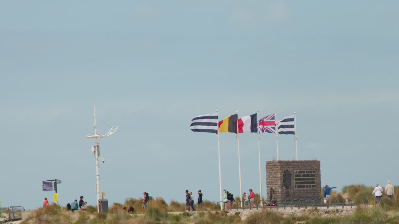 People stroll along coastal pathway by memorial monument, flags waving, under bright natural daylight