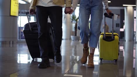 le immagini frontali ritagliate di una coppia di viaggiatori che trasportano i loro bagagli e si tengono per mano. camminano sulla sala del terminal. le persone si preparano ad imbarcarsi e partire con le valigie in rotolamento. slow motion