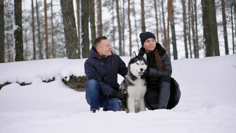 a man and a woman sitting hugging a dog siberian husky in the winter forest smiling and looking at each other and at the camera. slow motion happy family