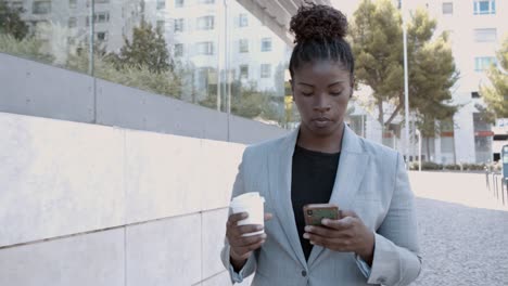 dolly shot of a focused african-american businesswoman walking outside, holding coffee and texting message on mobile phone